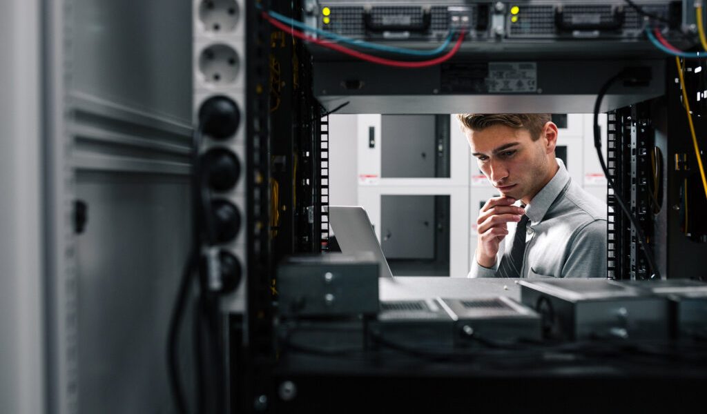 IT professional inspecting a server rack, looking at a laptop in a data center.