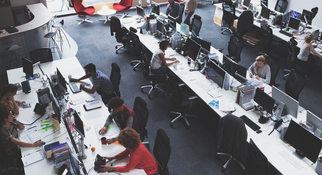 Oficina de planta abierta con escritorios largos y múltiples estaciones de trabajo informáticas; los empleados trabajan en sus ordenadores en medio de grandes ventanas y sillones rojos dispersos al fondo.