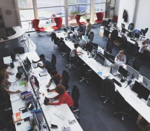 Oficina de planta abierta con escritorios largos y múltiples estaciones de trabajo informáticas; los empleados trabajan en sus ordenadores en medio de grandes ventanas y sillones rojos dispersos al fondo.