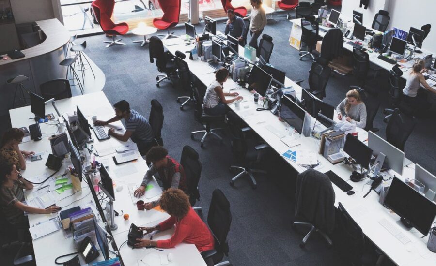 Oficina de planta abierta con escritorios largos y múltiples estaciones de trabajo informáticas; los empleados trabajan en sus ordenadores en medio de grandes ventanas y sillones rojos dispersos al fondo.