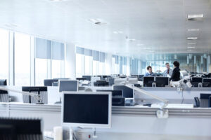 Oficina de planta abierta con filas de escritorios y monitores de computadora; un pequeño grupo de tres compañeros de trabajo charlando al fondo.