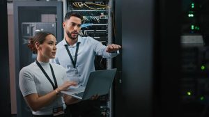 Dos profesionales de TI en un centro de datos revisan un ordenador portátil junto a los racks de servidores, y uno de ellos señala con un gesto hacia los equipos.