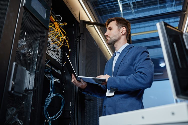 An IT professional in a blue suit inspecting server racks with a laptop in a data centre