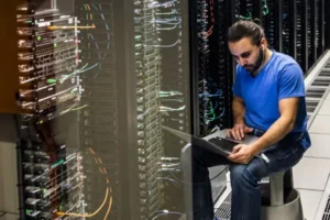 Tech worker in a blue shirt sits beside server racks, using a laptop in a data center to troubleshoot.